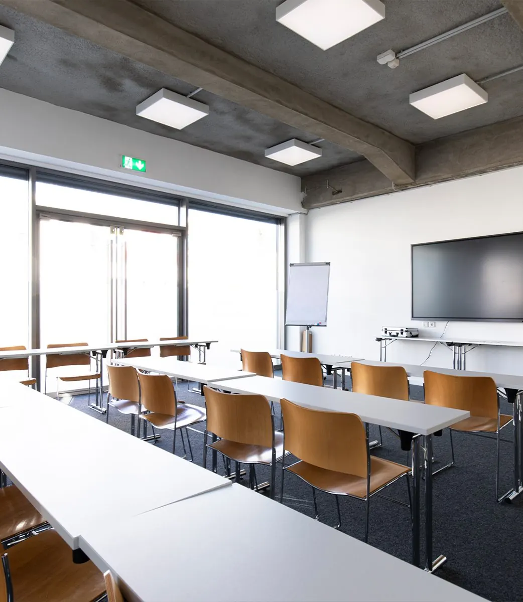 Brain Room: Tischreihen mit Stühlen, Flipchart und Bildschirm vor großen Fenstern
