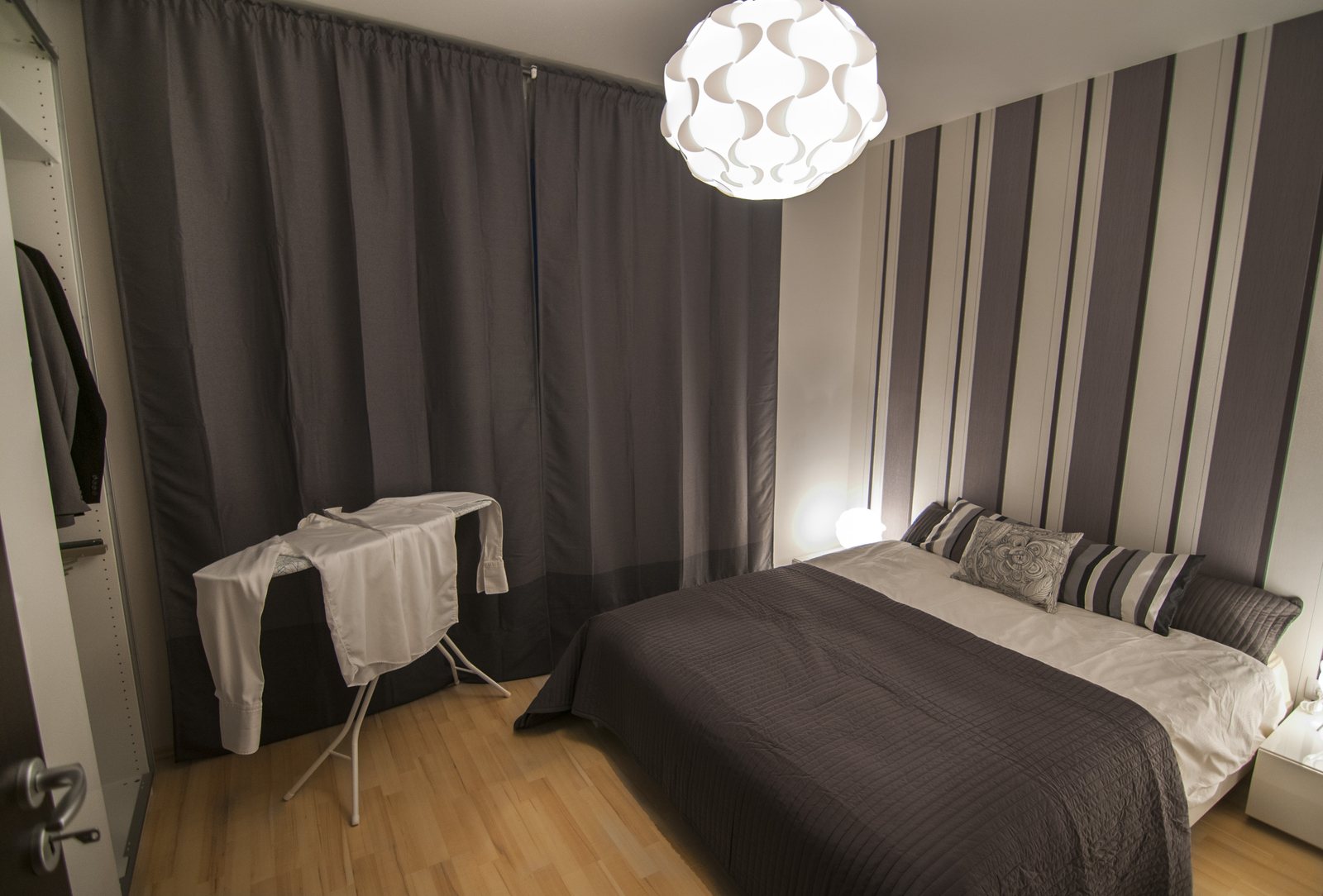Schlafzimmer einer Wohnung mit Doppelbett, gestreifter Tapete und Bügelbrett